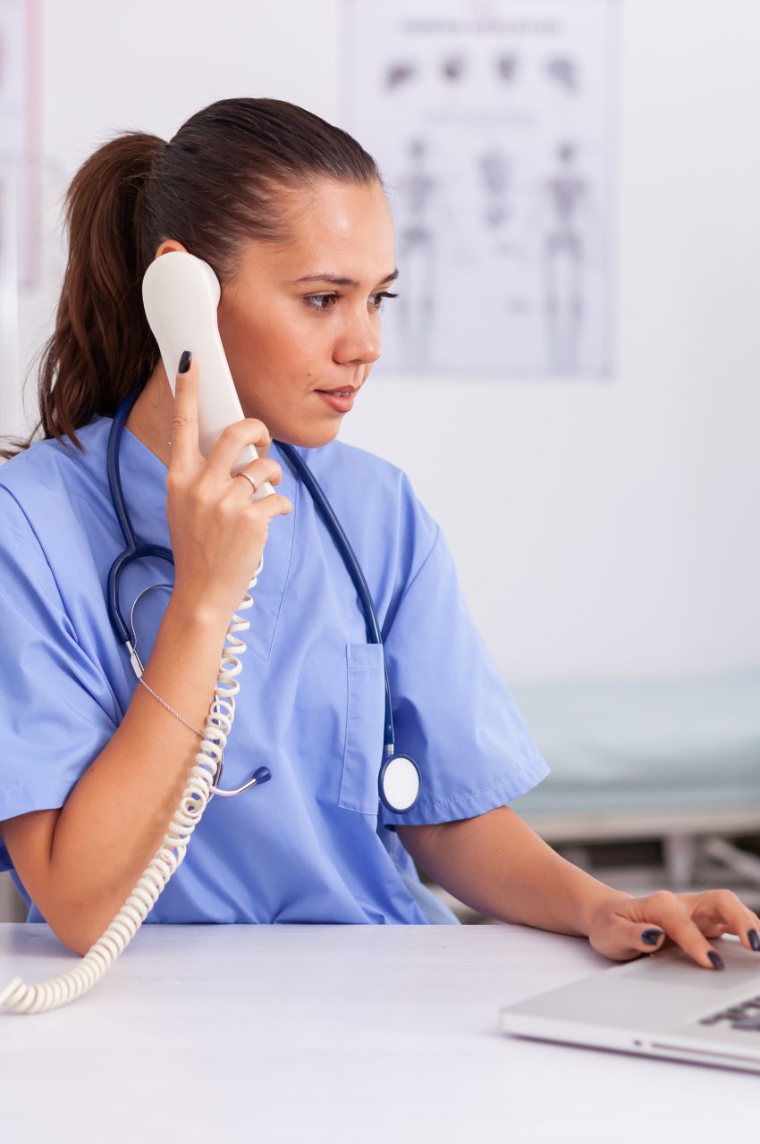 Medical practitioner answering phone calls and scheduling appointments in hospital office. Health care physician sitting at desk using computer in modern clinic looking at monitor.
