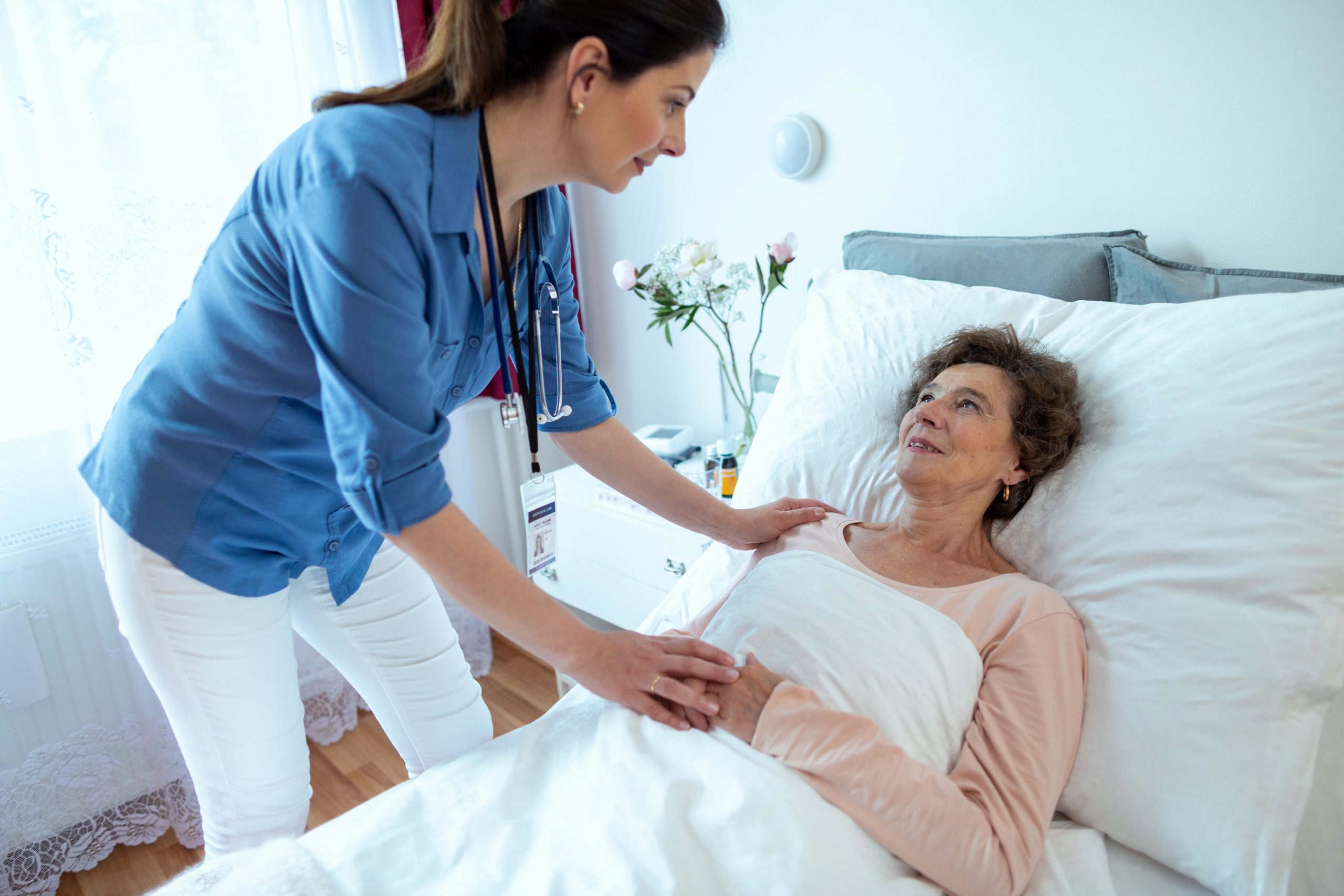 Nurse Leaning Over Elderly Woman Lying in Hospital Bed. Senior Female Patient Looking at Home Caregiver Holding Her Hand.